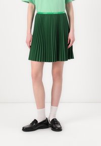 Grønn plissert skjørt med en tettsittende midje som har et merke-logo. Bært med svarte loafers og hvite sokker. Glatt materiale og middels lengde.