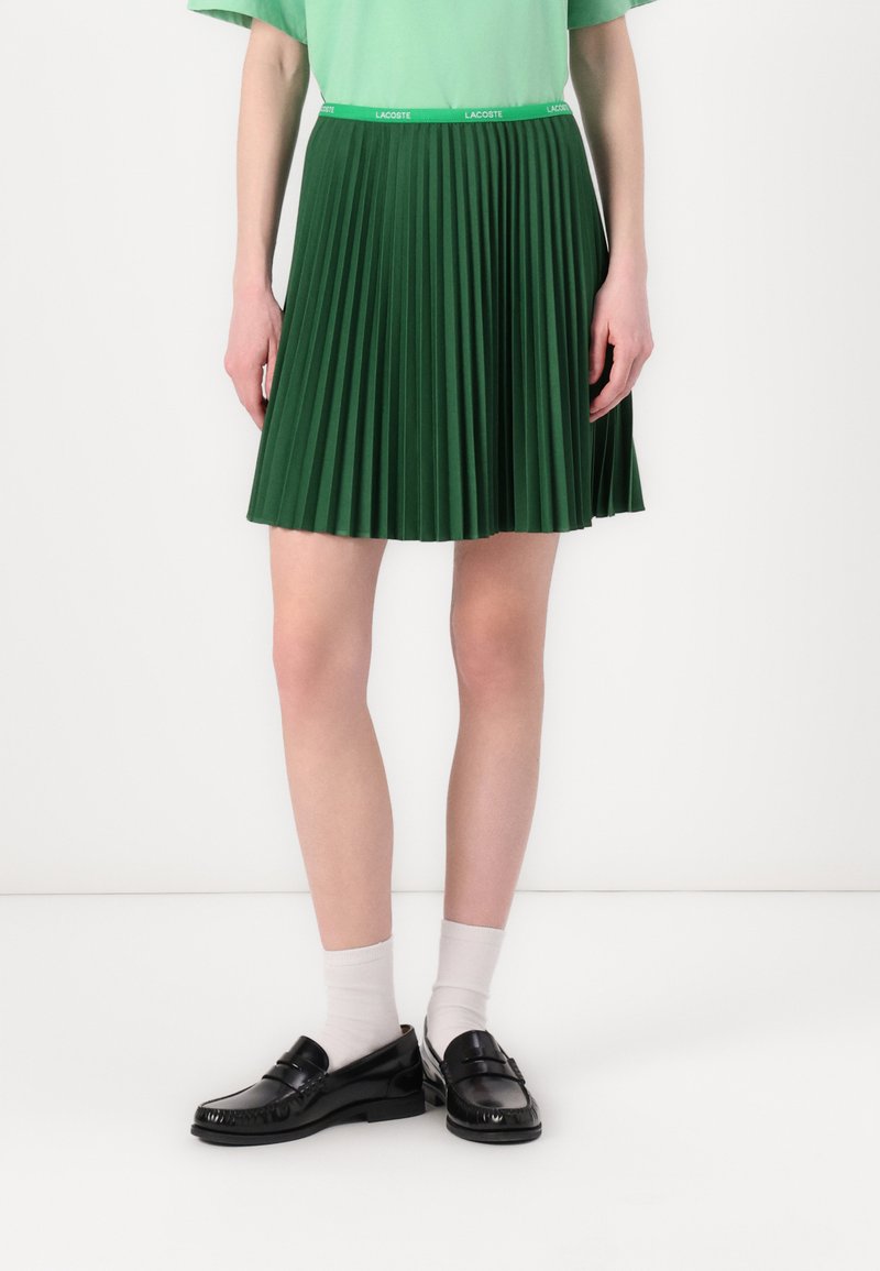 Grønn plissert skjørt med en tettsittende midje som har et merke-logo. Bært med svarte loafers og hvite sokker. Glatt materiale og middels lengde.