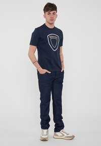 Marineblå T-shirt med et stort gråt skjoldlogo, parret med mørke denimjeans og sneakers med beige accenter. Enkel, tætsiddende design.