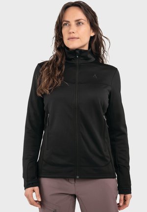 Frau trägt eine schwarze Jacke mit Reißverschluss und hohem Kragen sowie Reißverschlusstaschen, kombiniert mit mauvefarbigen Hosen, leicht nach rechts gewandt.