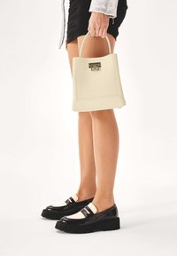 Borsa a tracolla in pelle crema con una forma strutturata, caratterizzata da una chiusura metallica decorativa. Scarpe con plateau nere lucide con accenti bianco sporco.