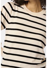 Femme portant un pull crème à rayures noires horizontales et épaules bouffantes, montrant le haut du torse et le cou.