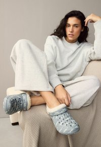 Lichtblauwe klompen met witte accenten en een voering van bont, voorzien van ventilatiegaten, gedragen op de voeten van een model, gecombineerd met grijze loungewear.