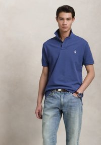 Νεαρός άνδρας στέκεται με το χέρι στην τσέπη, φορώντας μπλε πόλο μπλούζα με ανασηκωμένο γιακά και ανοιχτόχρωμα μπλε τζιν, μπροστά σε απλό φόντο.