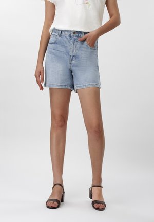 Nainen pukeutuneena vaaleansinisiin farkkushortseihin ja mustiin avokkaisiin, toinen käsi taskussa, seisomassa yksinkertaisen taustan edessä.