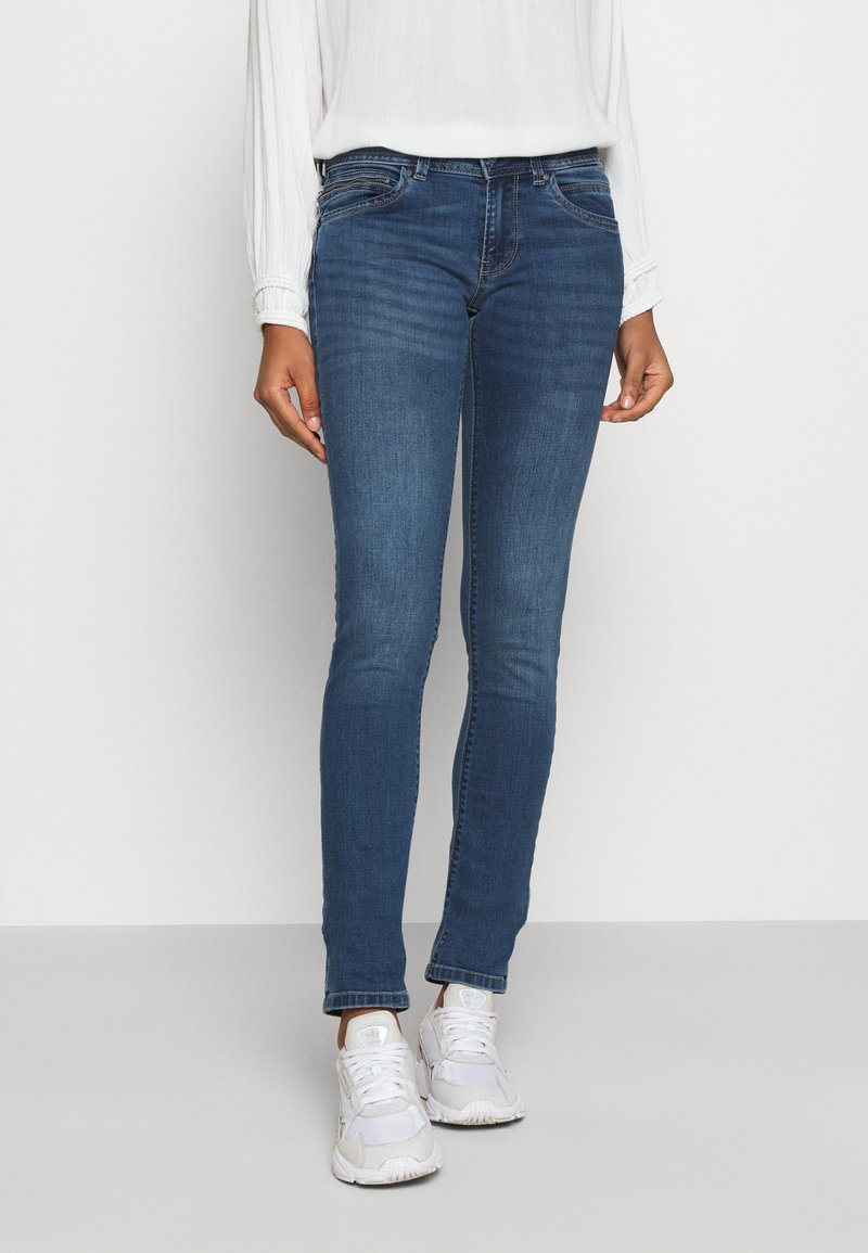 Femme portant un jean slim bleu, un haut blanc à manches longues et des baskets blanches, debout devant un fond clair uni.