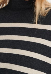 Pull à col roulé à rayures horizontales noires et beiges porté par une personne aux longs cheveux blonds visibles sur le côté droit.