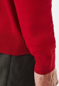 Czerwony sweter dzianinowy z żebrowanym wykończeniem u dołu i przy mankietach. Gładka faktura, dopasowany krój oraz subtelne detale przeszyć. Noszony z ciemnymi spodniami.
