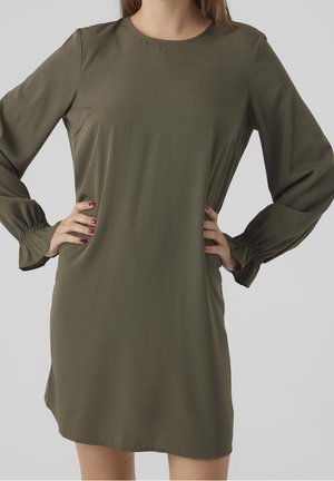 Vrouw draagt een olijfgroene jurk met lange mouwen en geplooide manchetten, handen in de zij, vooraanzicht tot halverwege de dij, tegen een effen achtergrond.