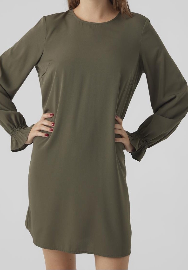 Femme portant une robe à manches longues vert olive avec poignets froncés, mains sur les hanches, vue de face jusqu'à mi-cuisse sur un fond uni.