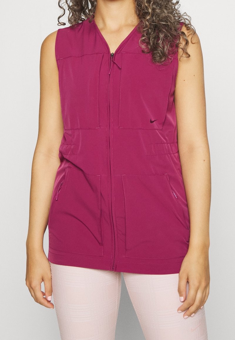 Personne portant un gilet sans manches magenta zippé avec des poches, associé à des leggings texturés rose clair, debout devant un fond uni.