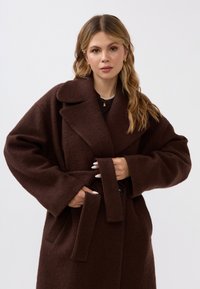 Brauner Wollmantel mit einem weiten Kragen, übergroßen Ärmeln und einem Bindegürtel. Verfügt über einen strukturierten Stoff und einen einzelnen Knopfverschluss.