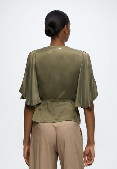 Femme aux cheveux foncés en chignon, portant un haut court olive à manches courtes avec détail d'épaule ouverte et un pantalon beige, dos au spectateur.