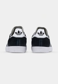 Svarte Adidas sneakers i semsket skinn sett bakfra, med hvite hælfaner som har svarte Adidas trefoil-logoer og hvite gummisåler.