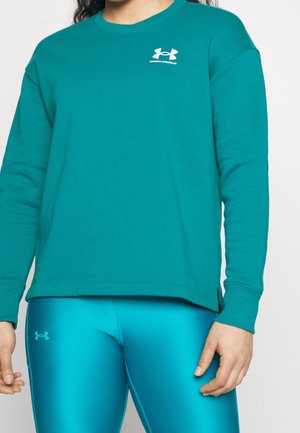 Osoba ubrana w turkusową bluzę Under Armour oraz pasujące turkusowe legginsy z logo na piersi i udzie, stojąca na jednolitym tle.