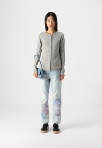 Szary wełniany kardigan z okrągłym dekoltem i guzikami, w zestawieniu z jasnoniebieskimi dżinsami z graficznymi naszywkami oraz czarnymi trampkami.