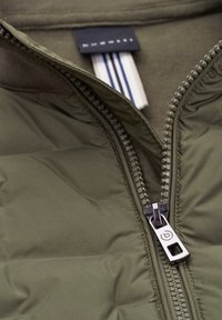 Olive green puffer jacket with a high collar, featuring a metal zipper and white striped label. Soft surface texture and quilted design.