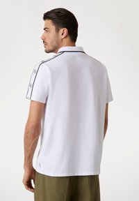 Camisa polo de manga corta blanca con detalles en negro y logotipo en las mangas. Tejido suave con un cuello clásico y pequeñas aberturas en los laterales.