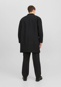 Svart ullkappa med rak skärning, med slagkrage och sidofickor, kombinerad med svarta byxor och skor.