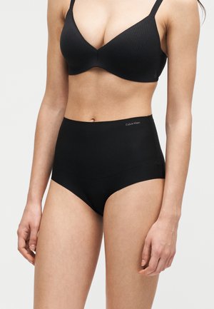 Femme portant un soutien-gorge noir sans coutures et un slip gainant taille haute avec un petit logo Calvin Klein sur la ceinture.