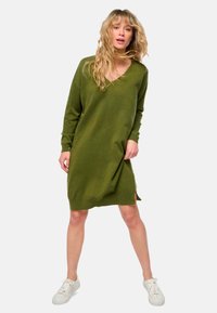 Femme aux cheveux blonds ondulés portant une robe pull V en jersey vert olive à manches longues et des baskets blanches, se tenant les jambes écartées.