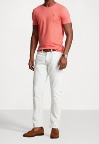 Koralowa koszulka z okrągłym dekoltem w połączeniu z białymi dżinsami, uzupełniona brązowym paskiem i brązowymi loafersami. Zegarek z skórzanym paskiem na nadgarstku.