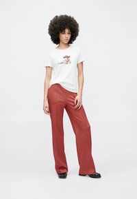 Weißes T-Shirt mit einem Tiermotiv; rote gemusterte Hose mit Rautendesign; Modell steht vor einfachem Hintergrund und trägt schwarze Schuhe.