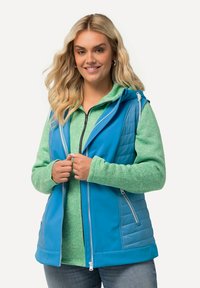 Gilet blu con pannelli texture, cappuccio e zip frontali, indossato sopra una felpa verde con macchie. Presenta una vestibilità rilassata e tasche laterali.