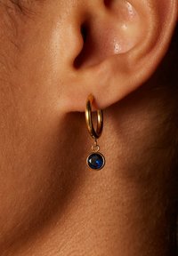 Gouden ringoorbel met een hangende blauwe facette-gemstone. De oorbel heeft een gladde afwerking en een glanzende goudtint.