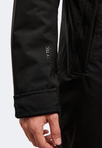 Gros plan sur la main d'une personne portant une veste imperméable Helly Tech noire avec un poignet réglable et un marquage visible sur la manche.