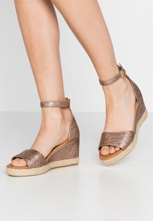 Pieds portant des sandales compensées couleur or rose avec des brides à la cheville et les orteils découverts sur un fond clair uni.