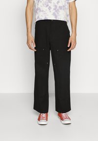 Breda svarta byxor med två framfickor. Tillverkade av ett slätt tyg, med silverfärgade detaljer. Matchas med ljusröda sneakers.