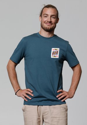 Mann mit Bart und zurückgebundenem Haar steht mit den Händen in der Hüfte, trägt ein türkisfarbenes Hemd mit einem Design der Könige der Herzen Spielkarte.