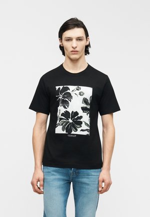 Νέος άνδρας που φοράει μαύρο μπλουζάκι με μεγάλο ασπρόμαυρο floral σχέδιο στο μπροστινό μέρος και ανοιχτό μπλε τζιν, στέκεται μπροστά σε απλό φόντο.