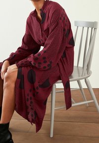 Abito bordeaux a maniche lunghe con pois neri e motivi astratti, dettaglio a bottoni al colletto e tessuto testurizzato. Modella seduta su una sedia.