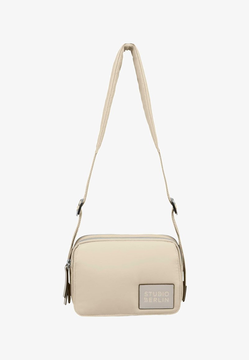Beige crossbody taske med en glat, polstret tekstur, lynlås lukning og et logo patch. Har en tynd, justerbar rem.