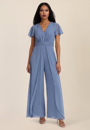 Frau steht, trägt einen ärmellosen, V-Ausschnitt, hellblauen Jumpsuit mit weitem Bein und taillierter Taille, kombiniert mit silberfarbenen offenen Absatzschuhen.