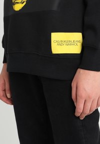 Černá mikina s velkým grafickým potiskem a žlutou nášivkou s nápisem "CALVIN KLEIN JEANS ANDY WARHOL", s žebrovanými manžetami a lemem.