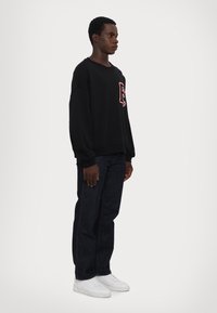 Zwarte sweatshirt met een rood logo, ontspannen pasvorm, geribbelde mouwen, gedragen met donkere jeans en witte sneakers, gefotografeerd tegen een effen achtergrond.