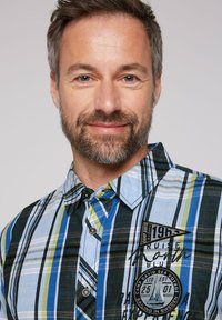 Camicia a maniche corte a quadri con strisce blu, verdi e nere. Presenta un colletto con bottoni e una toppa ricamata sul petto.