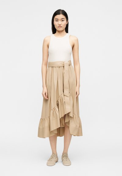 Γυναίκα που φοράει λευκό αμάνικο τοπ μέσα σε μπεζ φούστα τύπου wrap με βολάν και δεμένη ζώνη, σε συνδυασμό με μπεζ αθλητικά παπούτσια, στέκεται όρθια.