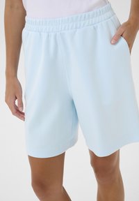Shorts de sport bleu clair avec un waistband élastique, poches latérales et tissu en mélange de coton lisse, présentant une coupe longueur genoux et un design minimaliste.