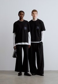 Čierne tričká s vreckom a grafickým detailom, kombinované s voľnými čiernymi nohavicami. Pozadie je neutrálne a minimalistické.