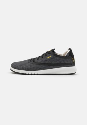 Zapatilla deportiva de punto en negro y gris con suela blanca, que presenta un diseño elegante, punta redonda y detalles en negro con una marca amarilla.