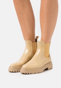 Bottes de pluie beiges brillantes à la cheville avec des semelles chunky portées sur des jambes nues sur un fond blanc.