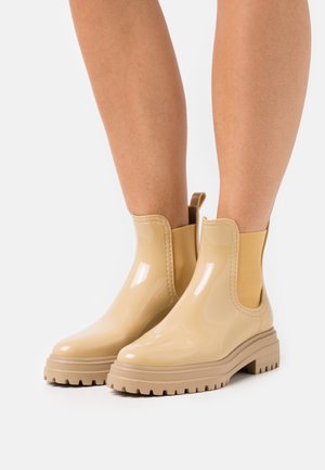 Bottes de pluie beiges brillantes à la cheville avec des semelles chunky portées sur des jambes nues sur un fond blanc.