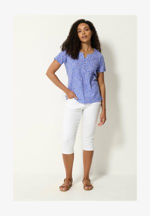 Mujer con pelo rizado que lleva una blusa azul de manga corta con flores, pantalones blancos recortados y sandalias marrones, de pie frente a un fondo blanco.