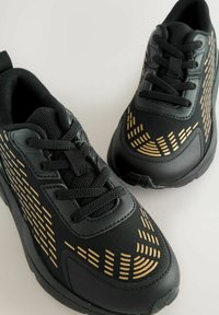 Ein Paar schwarze Sportschuhe mit goldenen geometrischen Mustern, Schnürdesign und gepolstertem Mesh-Futter auf einem hellen Hintergrund.