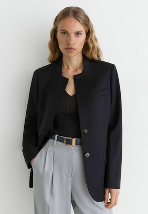 Vrouw met golvend blond haar draagt een zwart jasje met knopen, een zwarte top, grijze geplooide broek en een donkere riem met gouden gesp, staand in een neutrale houding.