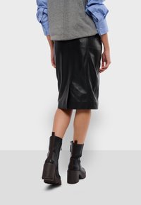 Jupe crayon en cuir noir, longueur genou, avec une fente à l'arrière ; portée avec un haut sans manches gris et des bottes noires à talons avec une semelle épaisse.